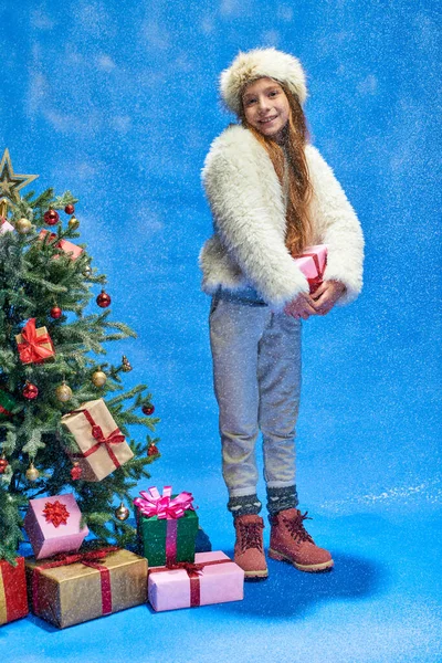 Cheerful girl in faux fur jacket and hat holding gift under falling snow near Christmas tree on blue — Stock Photo