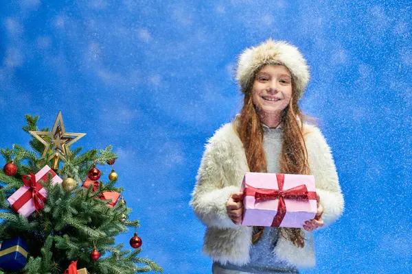 Joyful girl in faux fur jacket and hat holding gift under falling snow near Christmas tree on blue — Stock Photo