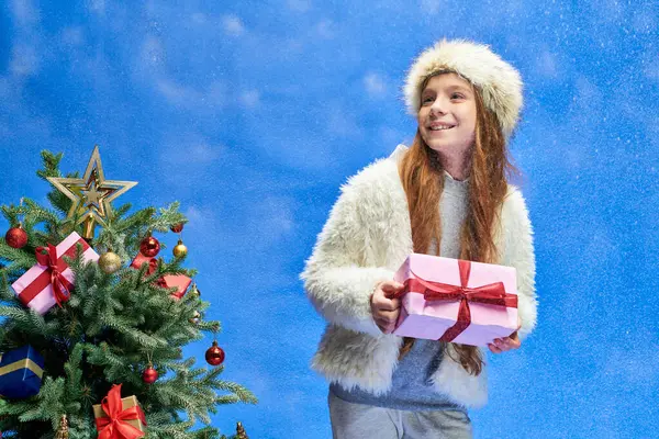 Excited girl in faux fur jacket and hat holding gift under falling snow near Christmas tree on blue — Stock Photo