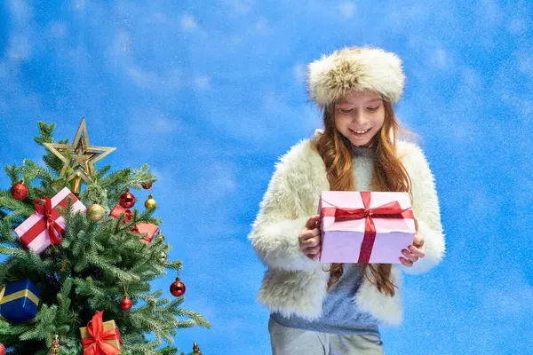 Chica excitada en chaqueta de piel sintética y sombrero mirando el regalo bajo la nieve que cae cerca del árbol de Navidad - foto de stock