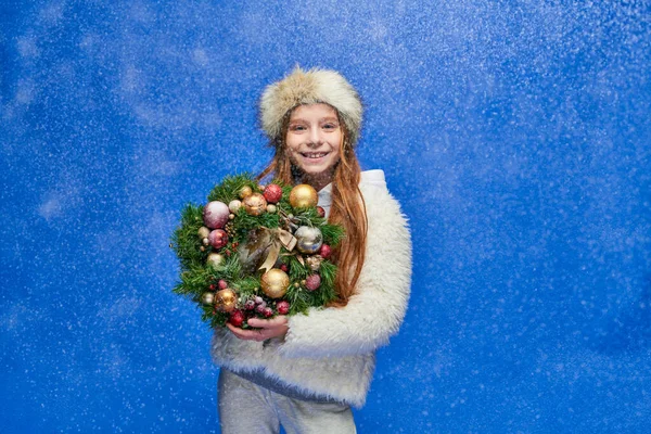 Happy girl in faux fur jacket and hat holding decorated Christmas wreath under falling snow on blue — Stock Photo