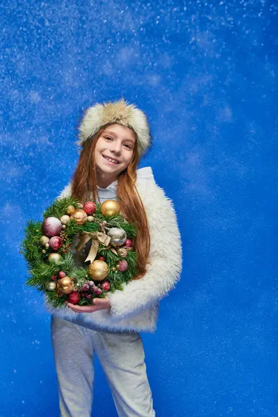 Positive girl in faux fur jacket and hat holding Christmas wreath under falling snow on blue — Stock Photo