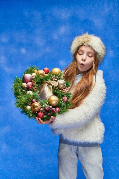 Emotional girl in faux fur jacket and hat holding Christmas wreath under falling snow on blue — Stock Photo