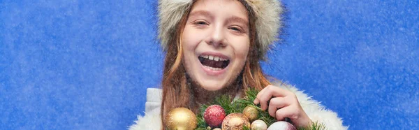 Excited kid in faux fur hat and jacket holding Christmas wreath under falling snow on blue, banner — Stock Photo