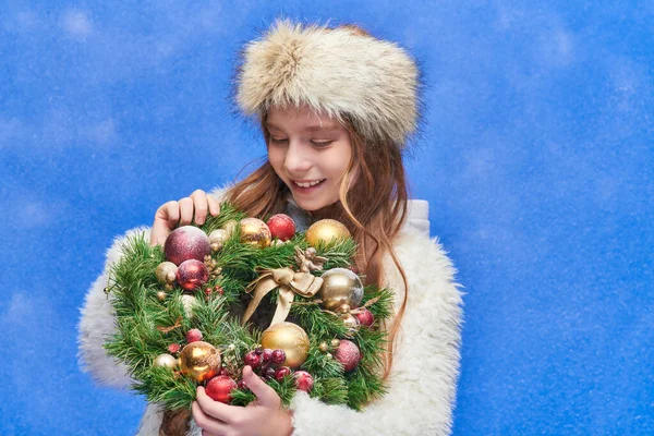 Happy child in faux fur hat and jacket looking at Christmas wreath under falling snow on blue — Stock Photo