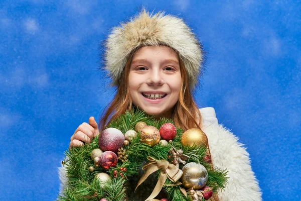 Temporada de alegría, niña preadolescente feliz celebración de la corona de Navidad bajo la caída de la nieve sobre fondo azul - foto de stock