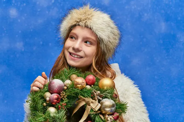 Temporada de alegría, niña preadolescente alegre celebración de la corona de Navidad bajo la nieve que cae sobre fondo azul - foto de stock