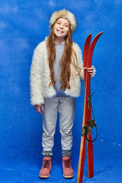 Cheerful preteen girl in winter faux fur jacket and hat holding red skis on turquoise background — Stock Photo