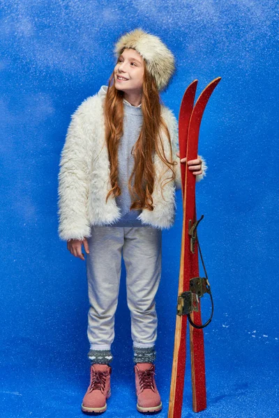Positive preteen girl in winter faux fur jacket and hat holding red skis on turquoise background — Stock Photo