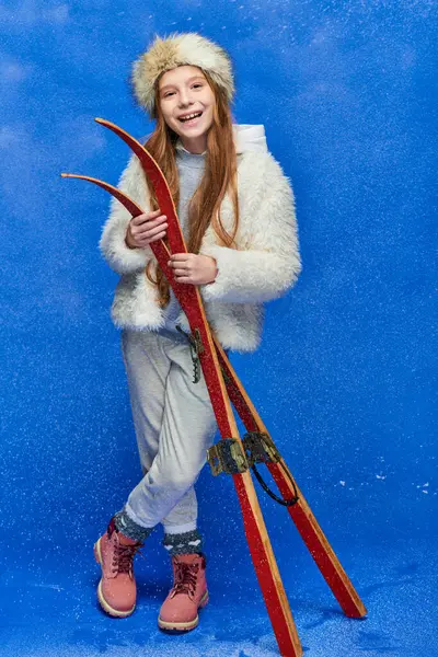 Smiling preteen girl in winter faux fur jacket and hat holding red skis on turquoise background — Stock Photo