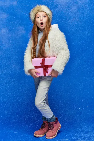 Winter joy, shocked preteen girl in faux fur jacket and hat holding gift box on turquoise background — Stock Photo