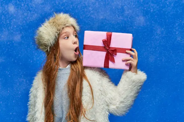Winter holidays, shocked girl in faux fur jacket and hat holding gift box on turquoise background — Stock Photo