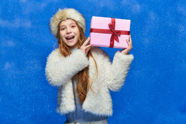 Vacances d'hiver, fille gaie en fausse veste de fourrure et chapeau tenant boîte cadeau sur fond turquoise — Photo de stock