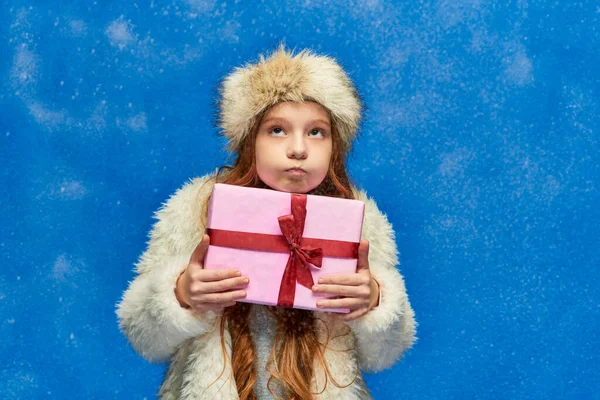 Vacances d'hiver, fille en fausse veste de fourrure et chapeau joues gonflantes et tenant boîte cadeau sur turquoise — Photo de stock
