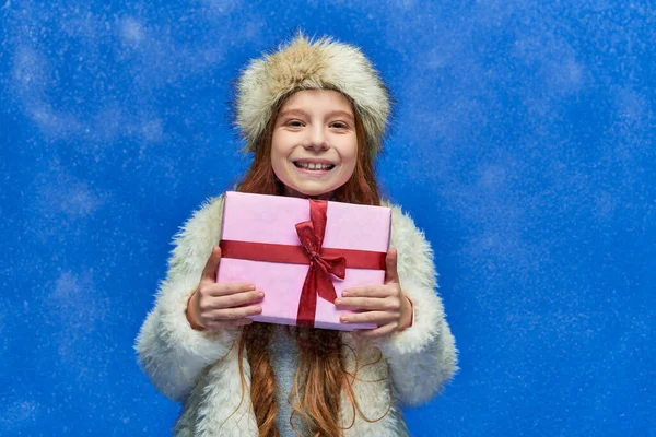 Vacances d'hiver, fille heureuse en fausse veste de fourrure et chapeau tenant boîte cadeau enveloppé sur turquoise — Photo de stock