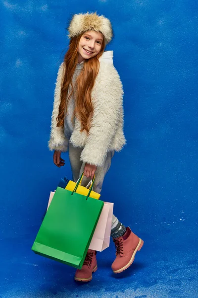 Winter shopping, happy girl in faux fur jacket and hat holding shopping bags on turquoise, snow — Stock Photo