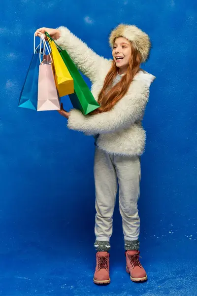 Compras de vacaciones, niño alegre en chaqueta de piel sintética y sombrero sosteniendo bolsas de compras en turquesa, nieve - foto de stock