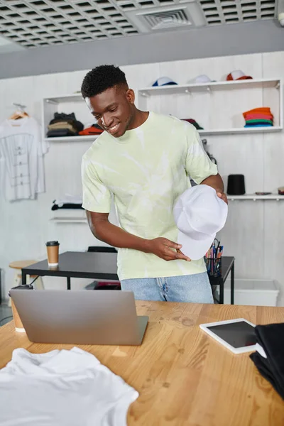 Happy african american designer showing white cap during video call on laptop in print studio — Stock Photo