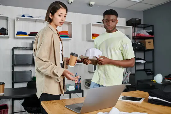 Designers multiétnicos com tampa branca e copo de papel olhando para laptop na mesa de trabalho em estúdio de impressão — Fotografia de Stock