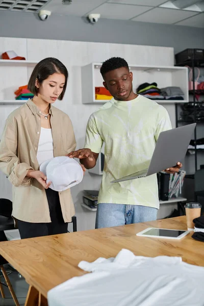 Multiethnic fashion designers with laptop and white cap discussing new project in print studio — Stock Photo