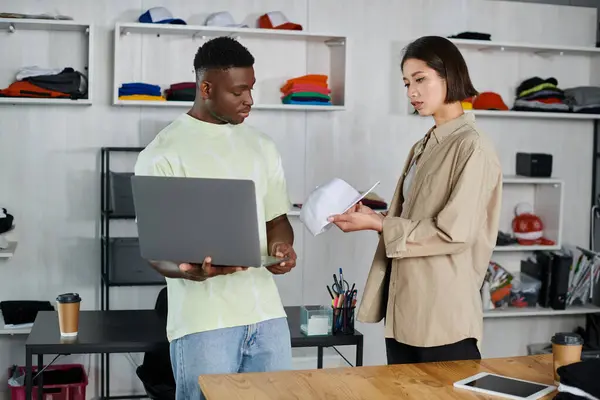 Asian woman showing white cap to african american colleague with laptop in clothing design atelier — Stock Photo
