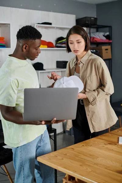 Jóvenes emprendedores interracial con gorra blanca y portátil discutiendo nueva startup en estudio de impresión - foto de stock