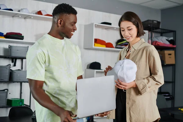 Smiling asian woman holding white cap near african american colleague with laptop in print studio — Stock Photo