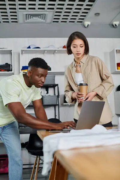 Inspirado homem americano africano usando laptop perto mulher asiática com copo de papel em estúdio de design de roupas — Fotografia de Stock