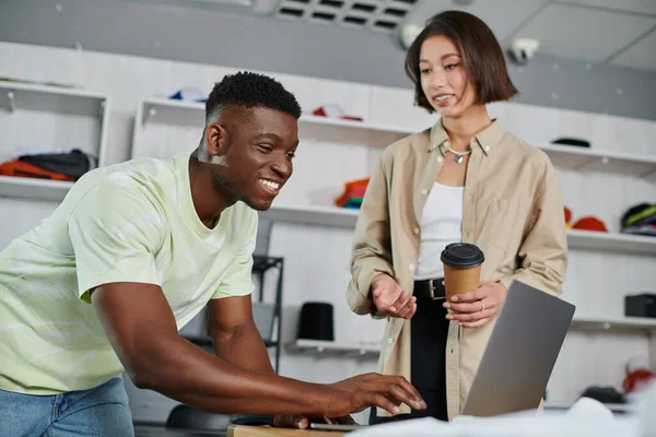 Cheerful african american man using laptop near asian colleague with coffee to go in print studio — Stock Photo