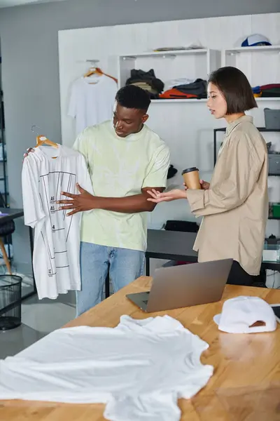 African american man showing white t-shirt with format sizes to asian colleague in print studio — Stock Photo