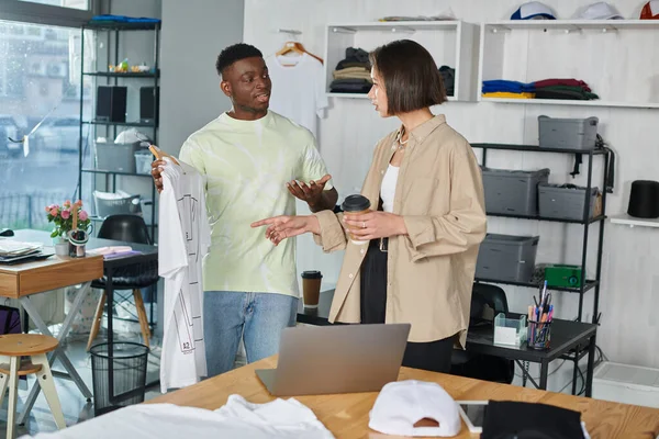 Multiethnic fashion designers sharing ideas near white t-shirt with format sizes in print studio — Stock Photo