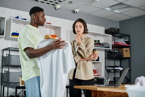 African american designer showing white t-shirt to thoughtful asian colleague in print studio — Stock Photo