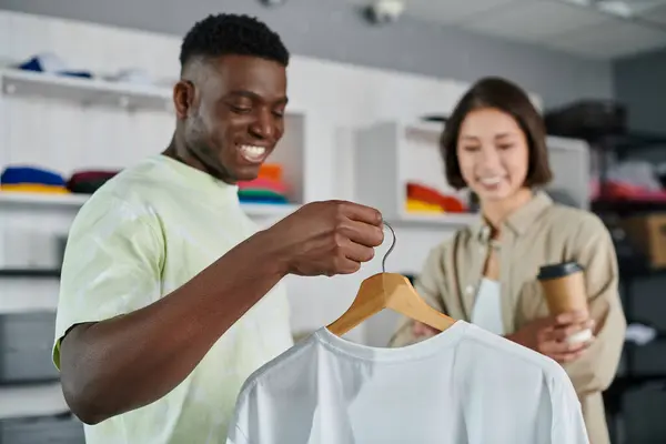 Веселый африканский американский дизайнер показывает белую футболку улыбающемуся азиатскому коллеге в типографии — стоковое фото