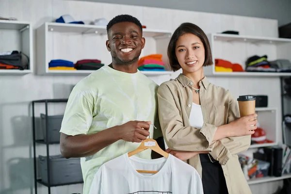 Joyful multiethnic designers with white t-shirt and coffee to go looking at camera in print studio — Stock Photo