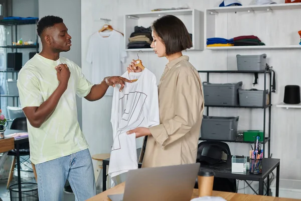 Africano americano diseñador apuntando a blanco camiseta cerca asiático colega en ropa impresión estudio - foto de stock