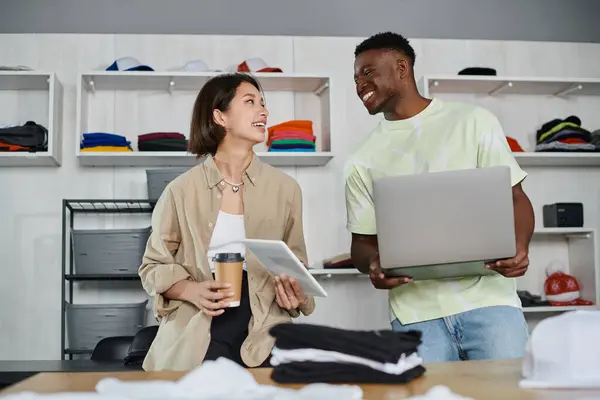 Joyful interracial designers with devices and coffee to go talking near clothes in print studio — Stock Photo