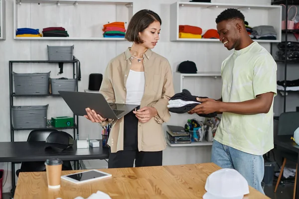 Happy african american designer holding clothing near asian colleague with laptop in print studio — Stock Photo