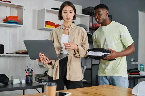 Asian designer pointing at laptop near african american colleagues holding clothes in print studio — Stock Photo