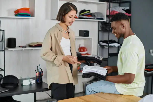 Smiling asian designer pointing at garments in hands of african american colleague in print atelier — Stock Photo