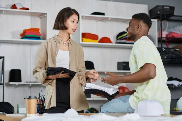 Jovens empreendedores inter-raciais segurando roupas e compartilhando ideias criativas no estúdio de impressão, trabalho em equipe — Fotografia de Stock