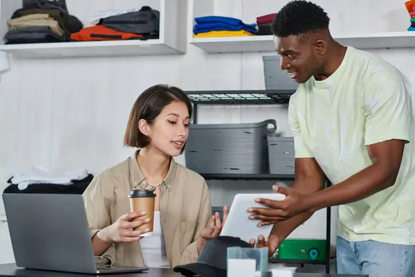 Happy african american designer showing digital tablet to asian woman with coffee to go near laptop — Stock Photo