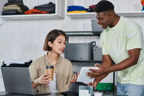 Feliz africano americano diseñador mostrando digital tablet a asiático mujer con café a ir cerca de portátil - foto de stock