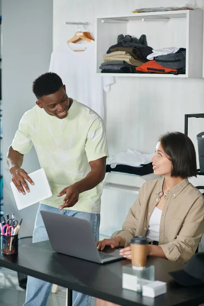 Designer americano africano com tablet digital conversando com mulher asiática perto de laptop em estúdio de impressão — Fotografia de Stock