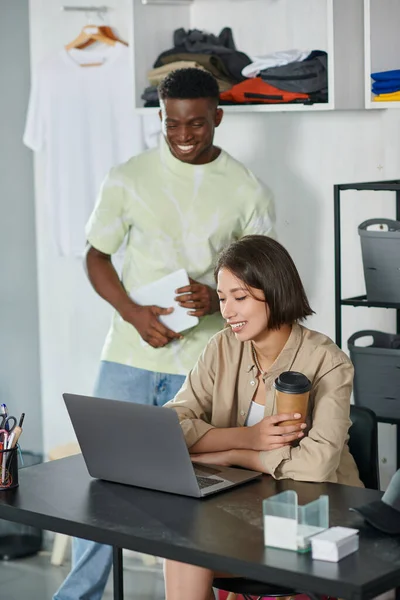 Щасливий азіатський дизайнер з паперовою чашкою, що працює на ноутбуці біля афроамериканського чоловіка з цифровим планшетом — стокове фото