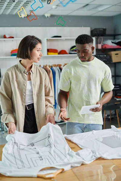 Jovem interracial pequenos empresários falando perto de camisetas com tamanhos de formato em estúdio de impressão — Fotografia de Stock