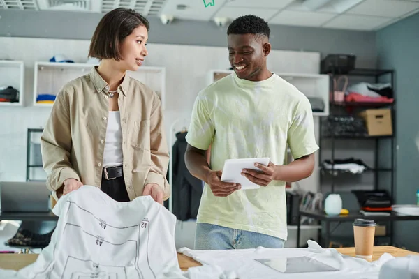 Young and creative interracial entrepreneurs talking near t-shirts with format sizes in print studio — Stock Photo