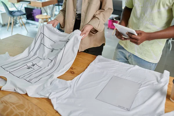 Vista cortada de designers de moda multiétnicos perto de camisetas brancas com tamanhos de formato em estúdio de impressão — Fotografia de Stock