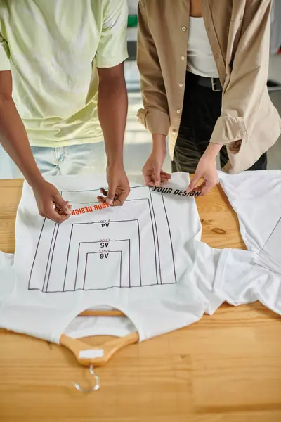 Vista cortada de designers inter-raciais segurando modelos de texto perto de t-shirts com tamanhos de formato — Fotografia de Stock