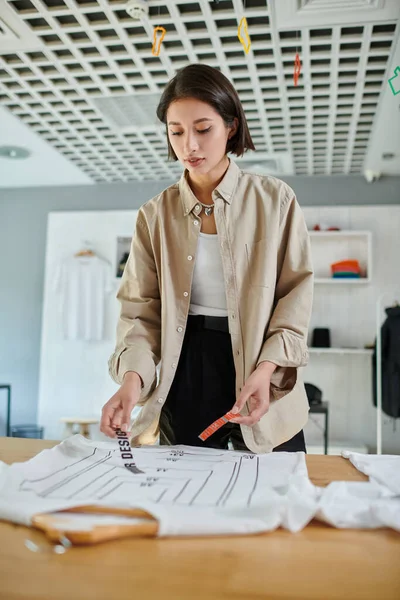 Jeune créatrice asiatique tenant le modèle de texte près de vêtements avec des tailles de format en studio d'impression — Photo de stock