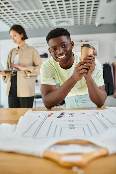 Щасливий афроамериканський дизайнер з паперовою чашкою дивиться на камеру біля азіатського колеги з ноутбуком — стокове фото